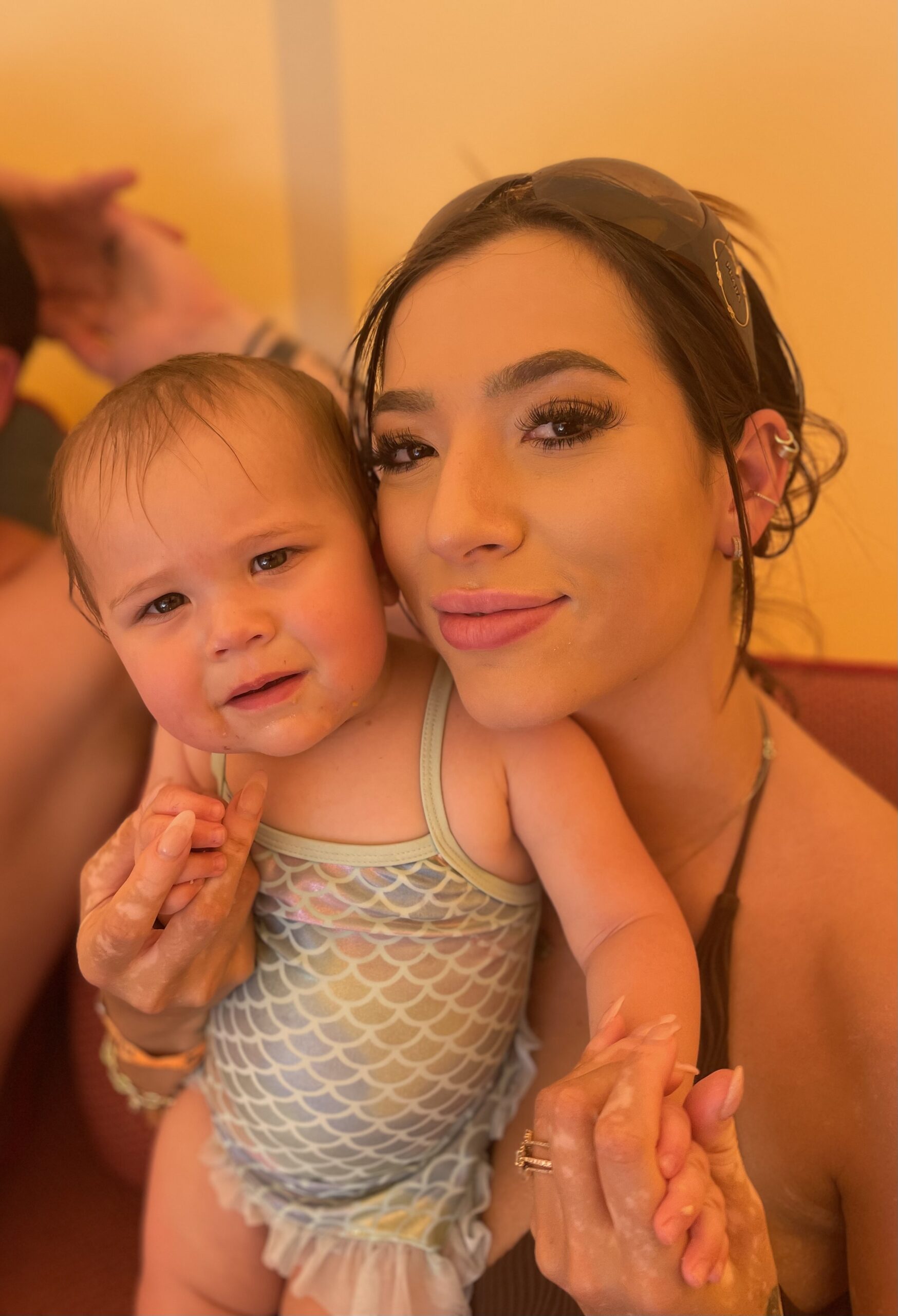 Growth Architect Beate Chelette's daughter Gina is holding her daughter in a light blue outfit. Both are looking at the camera and smiling, radiating warmth and happiness about this precious moment.