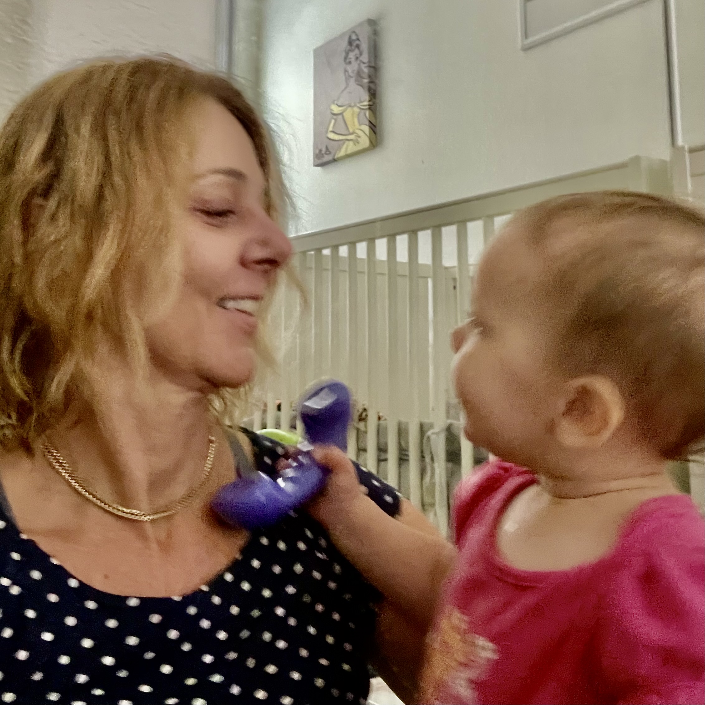 A woman in a polka dot shirt smiles while holding a baby dressed in pink. The baby looks at the woman, and they are inside a room with a crib in the background, sharing a tender moment about which memories are made.