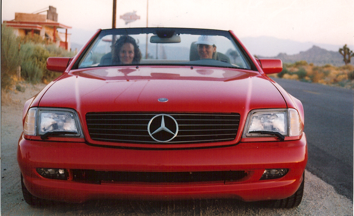 Beate Chelette in a red convertible Mercedes-Benz, parked on the roadside in a vast desert landscape with a building and mountains about in the background.