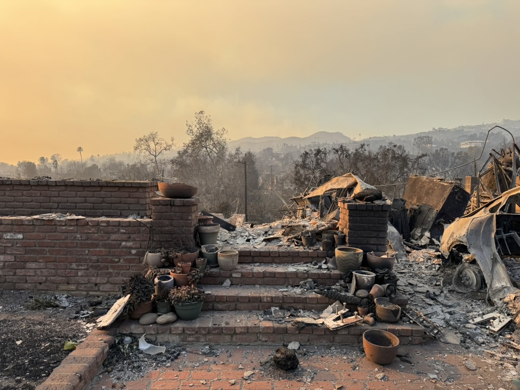 Amidst a burned landscape, a brick staircase leads to the charred remains of a house, with scattered pots in the foreground and a smoky sky looming overhead—a stark reminder to do better in 2025.