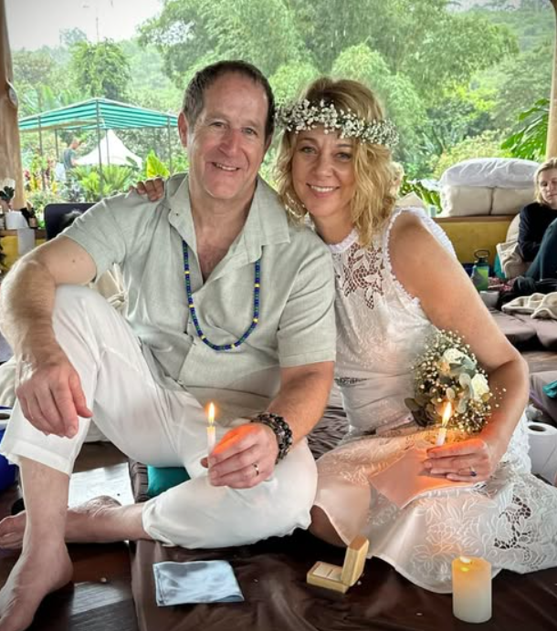 A couple sitting together, holding candles and dressed in white, symbolizes hope for a brighter future. The woman wears a floral crown and holds a bouquet, embodying the spirit to do better in 2025 amidst lush greenery in the background.