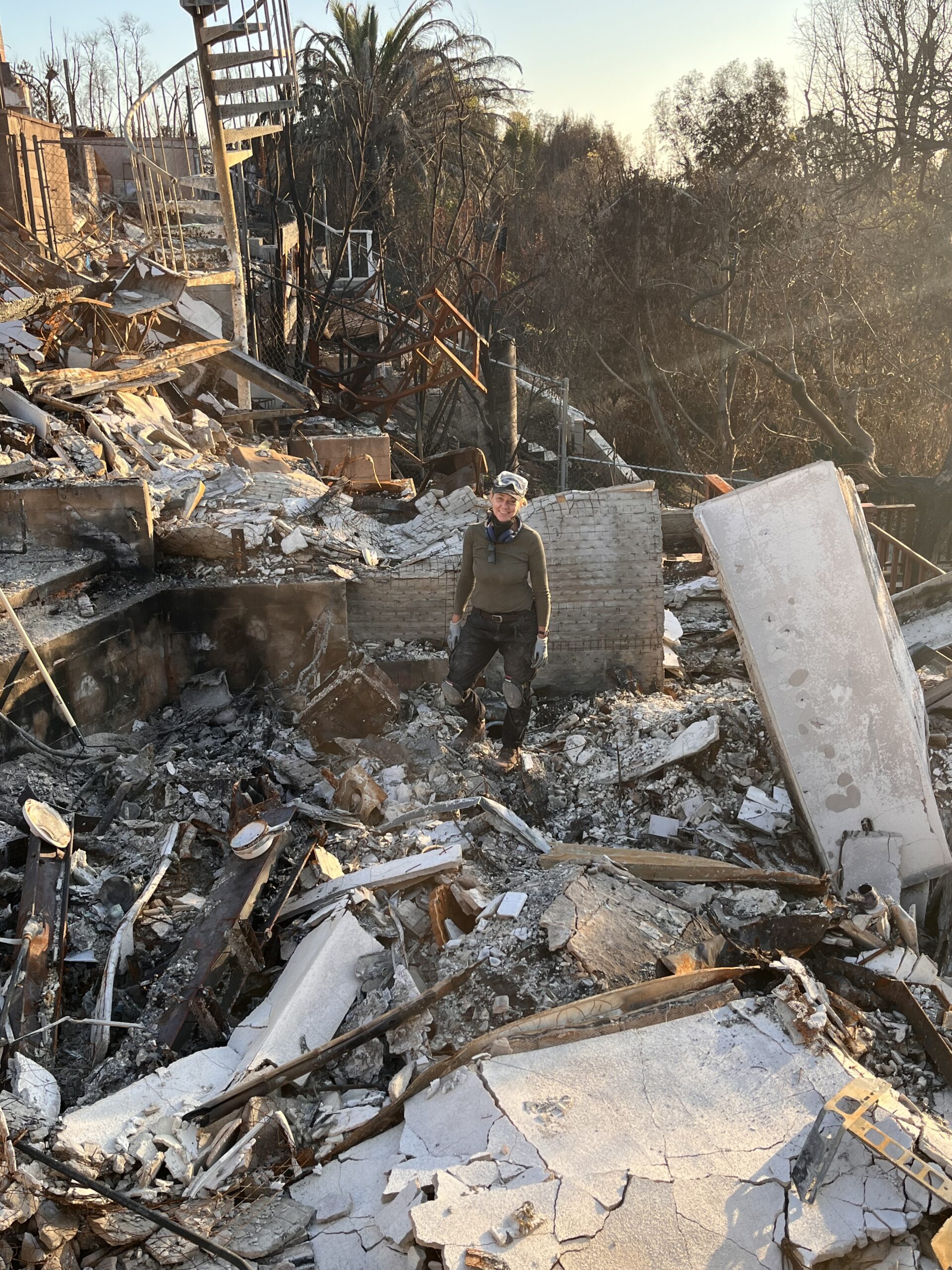 About: Beate Chelette wearing protective gear stands in the middle of debris and ruins from a burned-down building, surrounded by ash, rubble, and charred remains. About: Beate Chelette wearing protective gear stands in the middle of debris and ruins from a burned-down building, surrounded by ash, rubble, and charred remains.