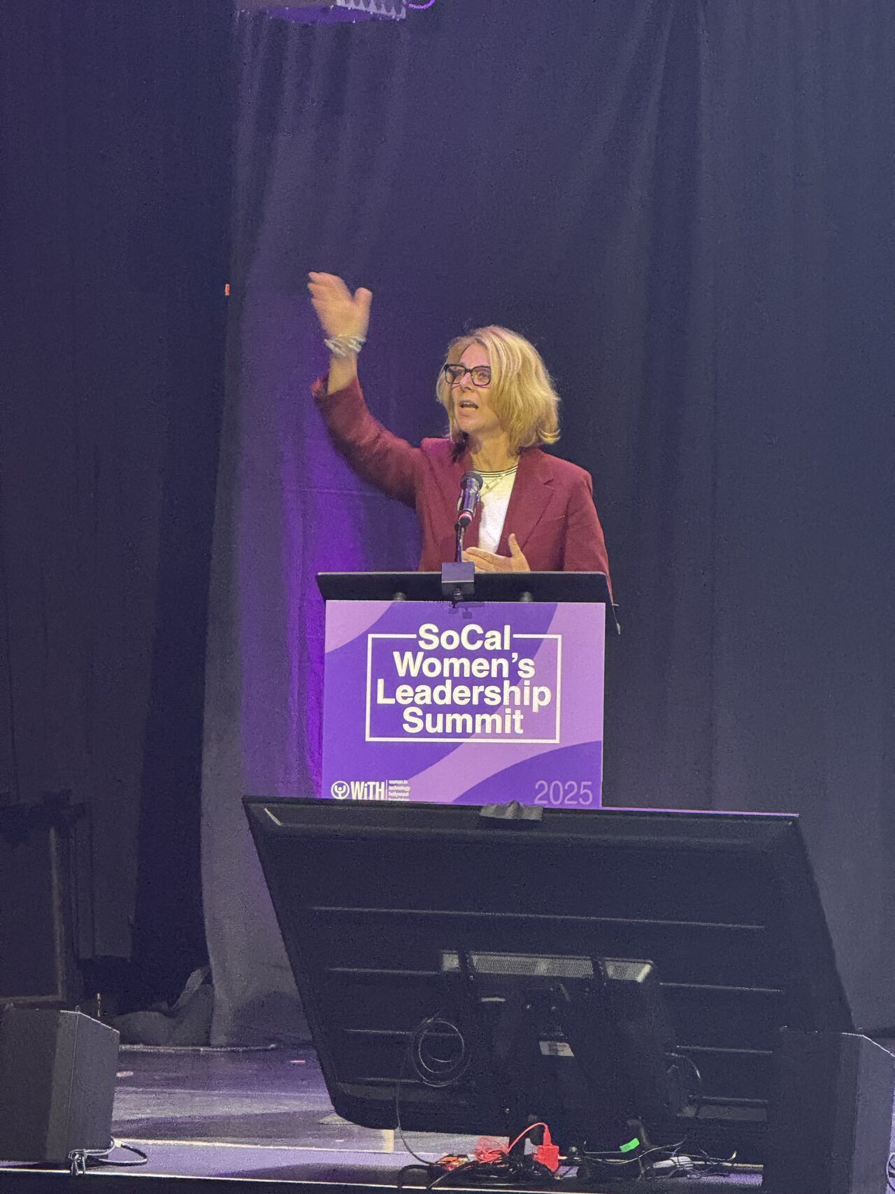 A woman speaks at a podium labeled "SoCal Women's Leadership Summit 2025," discussing how "AI Is Rising — How It Can Help You as a Small Business Owner" against a dark curtain backdrop.